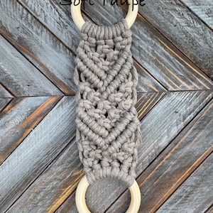 Pu&ograve; includere: Un arazzo in macram&egrave; con cordoncino di cotone color tortora tenue annodato in un motivo geometrico tra due anelli di legno liscio di colore chiaro. Il testo Soft Taupe &egrave; stampato in nero vicino all'anello di legno superiore.