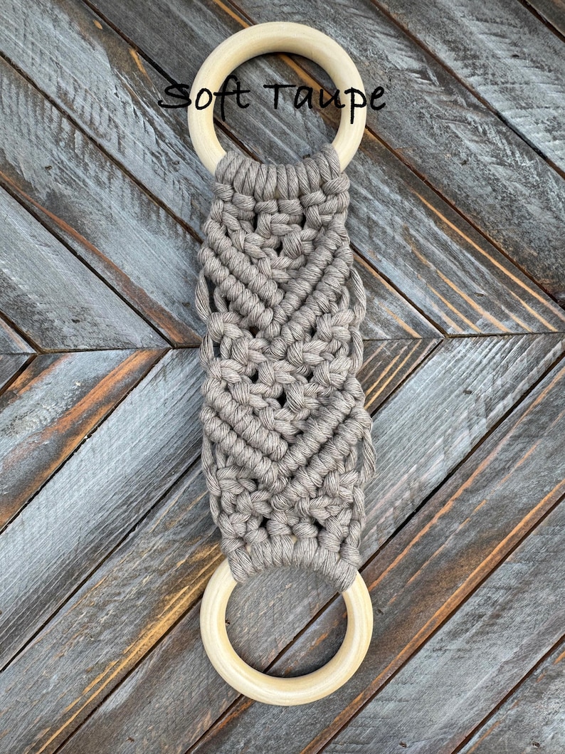 Pu&ograve; includere: Un arazzo in macram&egrave; con cordoncino di cotone color tortora tenue annodato in un motivo geometrico tra due anelli di legno liscio di colore chiaro. Il testo Soft Taupe &egrave; stampato in nero vicino all'anello di legno superiore.