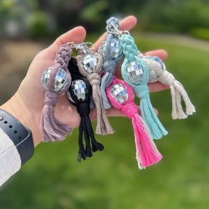 May include: A collection of colorful disco ball keychains. Each keychain features a small, mirrored disco ball attached to a braided cord in various colors, including pink, black, and blue. The keychains are held in a hand against a blurred green background.