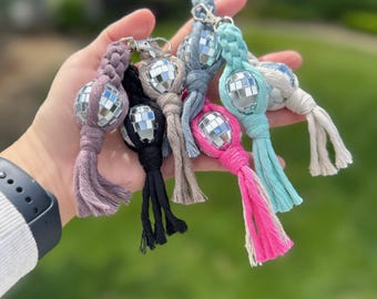 Può includere: Una collezione di portachiavi a sfera da discoteca colorati. Ogni portachiavi presenta una piccola sfera da discoteca a specchio attaccata a un cordoncino intrecciato in vari colori, tra cui rosa, nero e blu. I portachiavi sono tenuti in mano su uno sfondo verde sfocato.