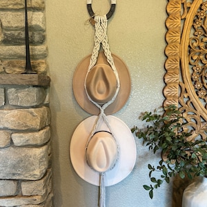 May include: A white macrame hat rack with a black horseshoe hanging on a wall. Three hats are hanging on the rack, one beige, one white, and one brown.