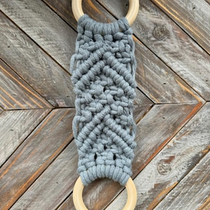 Pu&ograve; includere: Un arazzo in macram&egrave; con corda di cotone grigio annodata a motivo chevron, fissato a due anelli di legno chiaro e liscio nella parte superiore e inferiore. Il testo Cloudy Blue &egrave; stampato in nero vicino all'anello di legno superiore.