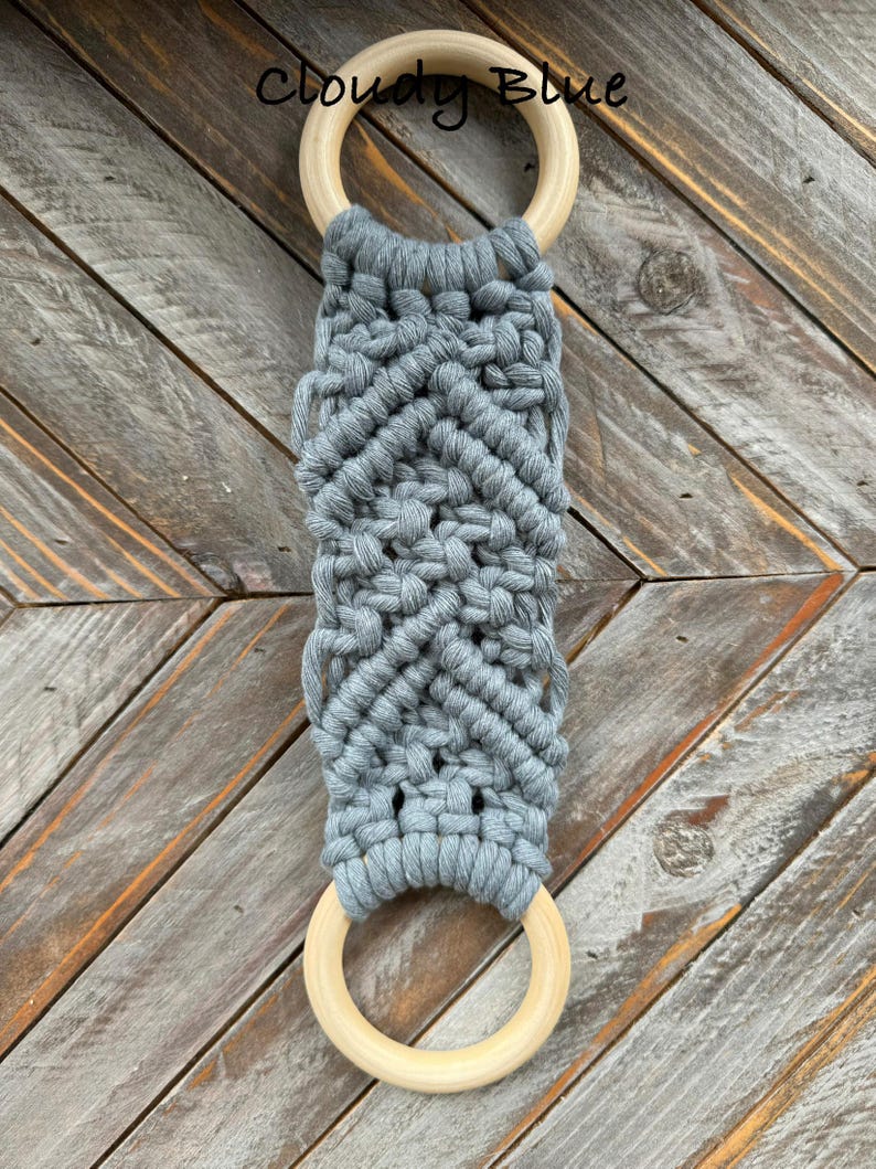 Pu&ograve; includere: Un arazzo in macram&egrave; con corda di cotone grigio annodata a motivo chevron, fissato a due anelli di legno chiaro e liscio nella parte superiore e inferiore. Il testo Cloudy Blue &egrave; stampato in nero vicino all'anello di legno superiore.