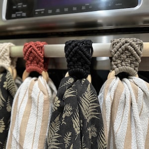 Pu&ograve; includere: Quattro asciugamani da cucina appesi con cime in macram&egrave; color crema, terracotta, nero e tortora. Ogni asciugamano &egrave; fissato a un anello di legno. I motivi includono felci dorate su fondo nero e strisce verticali bianche e beige.