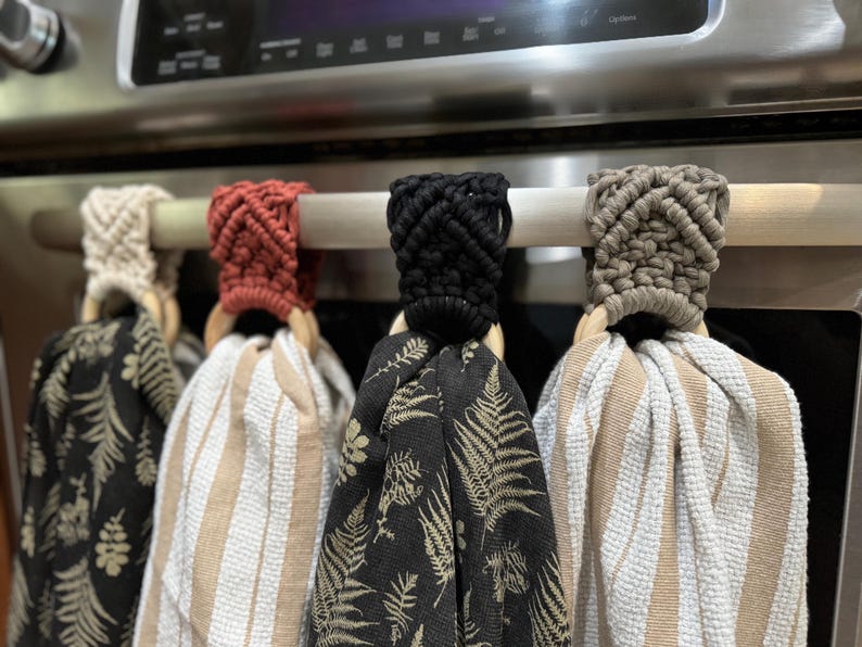 Pu&ograve; includere: Quattro asciugamani da cucina appesi con cime in macram&egrave; color crema, terracotta, nero e tortora. Ogni asciugamano &egrave; fissato a un anello di legno. I motivi includono felci dorate su fondo nero e strisce verticali bianche e beige.