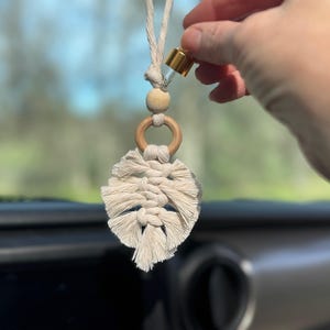May include: A white macrame leaf car air freshener with a wooden ring and bead. A hand is holding a small glass bottle with a gold lid, adding essential oil to the diffuser.