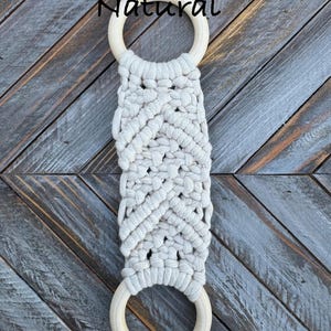 Pu&ograve; includere: Un arazzo in macram&egrave; con corda di cotone color bianco sporco annodata a motivo chevron, fissato a due anelli di legno liscio di colore chiaro in alto e in basso. La parola Natural &egrave; stampata in nero sopra l'anello superiore.