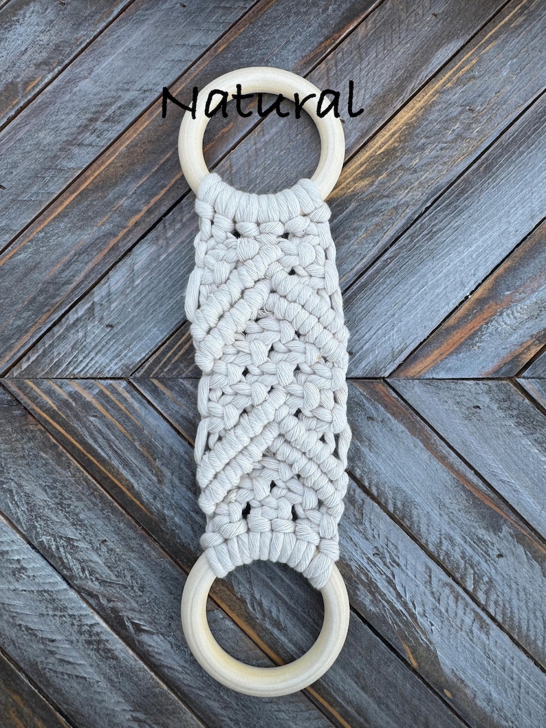 Pu&ograve; includere: Un arazzo in macram&egrave; con corda di cotone color bianco sporco annodata a motivo chevron, fissato a due anelli di legno liscio di colore chiaro in alto e in basso. La parola Natural &egrave; stampata in nero sopra l'anello superiore.