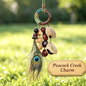 Puede incluir: Un adorno colgante con un anillo de madera oscura envuelto en hilo verde, decorado con una pluma de pavo real, semillas marrones y cáscaras de nuez texturizadas. Un letrero de madera clara muestra el texto Peacock Creek Charm.
