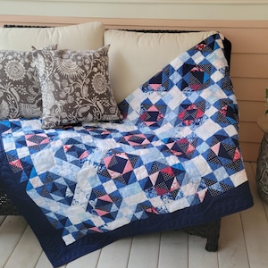 May include: A blue, pink, and white quilt with a geometric pattern is draped over a wicker chair with brown patterned pillows. The chair is on a porch with a wooden floor and a small table with a potted plant.