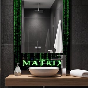 K&ouml;nnte beinhalten: Ein rechteckiger Wandspiegel mit einem schwarzen Rahmen, der mit gr&uuml;nen digitalen Codemustern und dem Wort MATRIX in einer leuchtend gr&uuml;nen Schrift am unteren Rand verziert ist. Der Spiegel reflektiert ein Badezimmer mit Duschkopf und Handtuch.