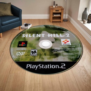 May include: A round rug featuring a close-up image of a PlayStation 2 game disc for Silent Hill 2. The disc is green and black with the game title, "Silent Hill 2", in white text. The game's rating, "Mature", is visible in the lower left corner. The game's publisher, "Konami", is visible in the upper right corner. The text "PlayStation 2" is visible in the lower center of the disc.