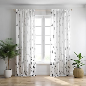 Puede incluir: Cortinas blancas y transparentes con un patrón de estrellas grises. Las cortinas cuelgan frente a una ventana, con una barra dorada. Las estrellas son de varios tamaños y están densamente agrupadas en la parte inferior.