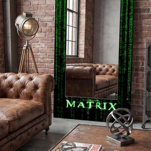 K&ouml;nnte beinhalten: Ein rechteckiger Standspiegel mit einem schwarzen Rahmen, der mit einem gr&uuml;nen digitalen Codemuster verziert ist und unten das Wort MATRIX in leuchtend gr&uuml;nen Buchstaben tr&auml;gt. Die Spiegelfl&auml;che reflektiert ein braunes Ledersofa in einem Raum.