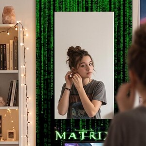 K&ouml;nnte beinhalten: Ein rechteckiger Wandspiegel mit einem schwarzen Rahmen, der mit einem gr&uuml;nen digitalen Codemuster verziert ist, das an Computer-Datenstr&ouml;me erinnert. Am unteren Rand des Rahmens steht das Wort MATRIX in fetten, leuchtend gr&uuml;nen Gro&szlig;buchstaben.