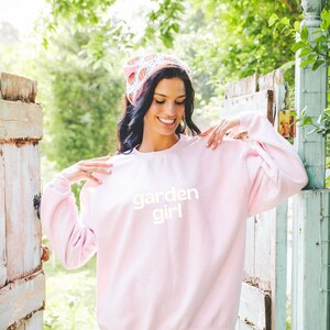 Puede incluir: Una sudadera rosa con el texto "garden girl" en letras blancas. La modelo lleva la sudadera con una diadema floral y pantalones cortos de mezclilla.