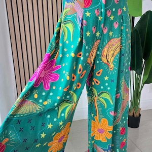 May include: Teal green wide-leg trousers with a bright tropical pattern. The print includes flowers, stars, bananas, and palm trees. The trousers have a button fastening and a pleated front.