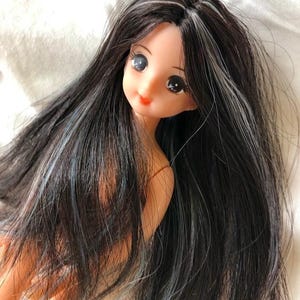 May include: A doll with long, dark hair with streaks of grey and blue. The doll has large, expressive eyes and a small, red mouth. The doll's skin is a pale peach colour. The doll is a collectible item.