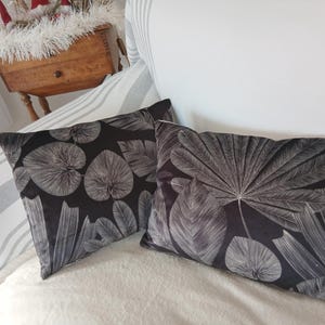 Könnte beinhalten: Zwei dekorative schwarze Kissen mit weißem Blättermuster. Die rechteckigen Kissen liegen auf einer weißen Oberfläche. Sie befinden sich auf einem weißen Sofa, mit einem gestreiften Kissen im Hintergrund.