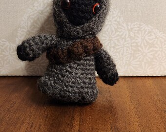May include: A small gray crocheted plush toy with brown accents and large orange eyes. The toy is sitting on a wooden surface.