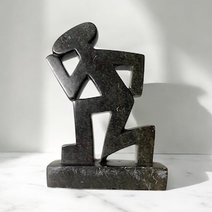 May include: A dark gray stone sculpture of a stylized figure in a kneeling position. The figure has a simple, geometric design with a rectangular base. The sculpture is made of a dark gray stone with visible texture and is displayed on a white surface.