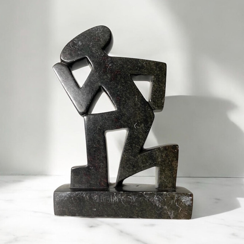 May include: A dark gray stone sculpture of a stylized figure in a kneeling position. The figure has a simple, geometric design with a rectangular base. The sculpture is made of a dark gray stone with visible texture and is displayed on a white surface.