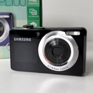 Könnte beinhalten: Eine schwarze Samsung Digitalkamera mit einem silber umrandeten Zoomobjektiv, das die Aufschrift 6.3-18.9mm 1:3.0-5.6 trägt. Die Kamera hat einen kleinen Blitz oben rechts und das Samsung-Logo auf der linken Seite, platziert vor der Originalverpackung.