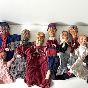 Puede incluir: Una colección de ocho marionetas de mano con rostros esculpidos y ropa de tela. Las figuras incluyen varios personajes con atuendos como una camisa azul con bufanda roja, una túnica granate, un vestido blanco y un traje negro, dispuestos en grupo.