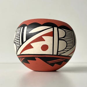 Op de afbeelding: Een kleine, ronde aardewerken kom in terracotta kleur met een zwart, wit en rood geometrisch ontwerp. De kom heeft een geschulpte zwarte rand en een herhalend patroon van lijnen, driehoeken en cirkels. Het ontwerp is symmetrisch en bedekt het gehele oppervlak.