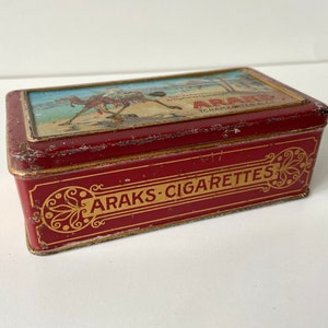 May include: A rectangular, vintage red tin with gold trim and lettering. The top features a colorful illustration of a person riding a camel. The words "ARAKS CIGARETTES" are displayed in gold lettering on the front of the tin.