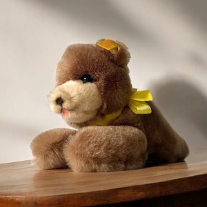 Könnte beinhalten: Ein brauner Plüschteddybär mit einer gelben Schleife. Der Bär liegt auf dem Bauch mit ausgestreckten Pfoten.