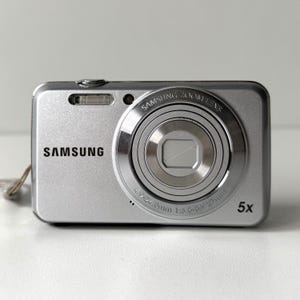 Może przedstawiać: Srebrny aparat cyfrowy Samsung z wyraźnym obiektywem Samsung Zoom Lens o specyfikacji 4.9-24.5mm 1:3.6-6.9 27mm. Na przedniej części kompaktowego urządzenia widnieje nazwa marki Samsung, lampa błyskowa, mały obiektyw oraz wskaźnik zoomu 5x.