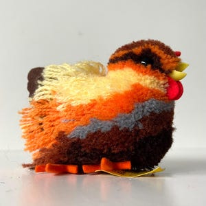 Op de afbeelding: Een klein, oranje, bruin en geel pluche kippen speelgoed met een rode kam en een gele snavel. De kip heeft een pluche textuur en staat op oranje poten.