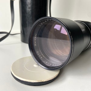 Puede incluir: Un objetivo de c&aacute;mara negro con el texto "Maroxar Auto Telephoto 135mm f/3.5" impreso en el lateral. El objetivo est&aacute; unido a un cuerpo de c&aacute;mara negro con una correa.