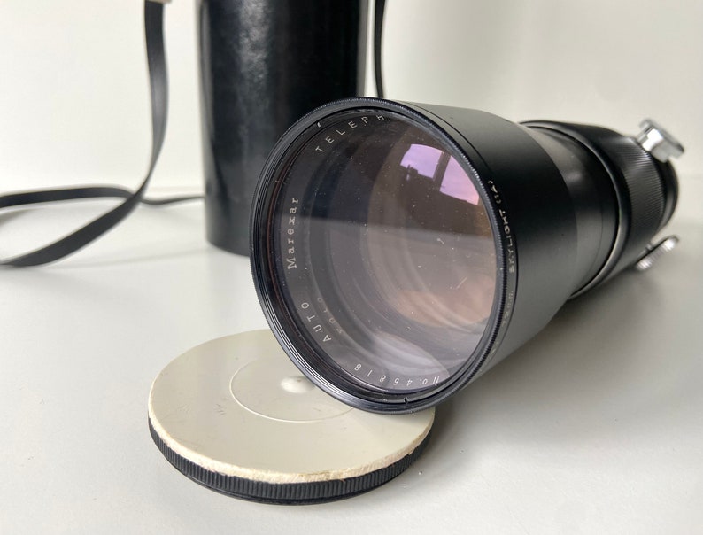 Puede incluir: Un objetivo de c&aacute;mara negro con el texto "Maroxar Auto Telephoto 135mm f/3.5" impreso en el lateral. El objetivo est&aacute; unido a un cuerpo de c&aacute;mara negro con una correa.