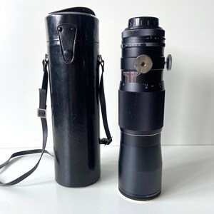 Puede incluir: Un objetivo de c&aacute;mara negro con una funda de transporte de cuero negro. El objetivo tiene una banda plateada alrededor del medio y un anillo plateado en la parte inferior. La funda tiene una correa.