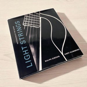 Op de afbeelding: Een boek getiteld "Light Strings: Impressions of the Guitar" door Ralph Gibson en Andy Summers. De cover is zwart met witte en lichtblauwe tekst en een gitaarafbeelding.