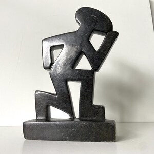 May include: A dark gray sculpture of a person kneeling, with one arm raised. The figure is stylized with geometric shapes and stands on a rectangular base. The sculpture has a matte finish.