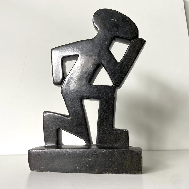 May include: A dark gray sculpture of a person kneeling, with one arm raised. The figure is stylized with geometric shapes and stands on a rectangular base. The sculpture has a matte finish.