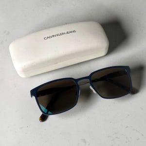 May include: A pair of dark blue rectangular sunglasses with dark tinted lenses and a white hard-shell protective case featuring the text CALVIN KLEIN JEANS printed in black on the top surface of the case lid.