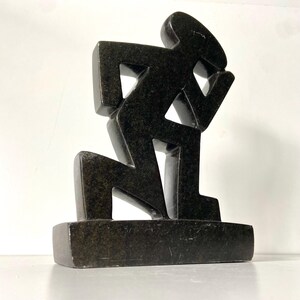May include: A dark, geometric sculpture of a kneeling figure. The sculpture is made of a dark, possibly stone-like material and is mounted on a rectangular base. The figure's form is angular and stylized.
