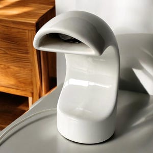 Op de afbeelding: Witte keramische tafellamp met een gebogen, golvend ontwerp. De lamp heeft een kap die aan één kant open is.