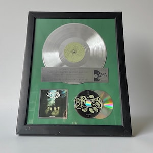 May include: Framed platinum-plated record and CDs. The frame has a black border and a green background. The record is silver with a yellow center. The CDs and album art are visible. Text on the plaque reads "Million Seller Award Status."