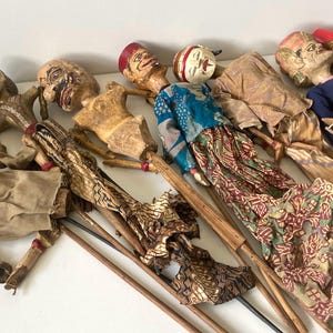 Op de afbeelding: Een verzameling van zeven Wayang Golek stokpoppen met gesneden houten hoofden, beschilderde gezichten en uitgebreide stoffen kleding. Elke pop is bevestigd aan een centrale houten bedieningsstok en toont diverse kleuren en patronen.