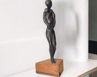May include: Bronze sculpture of a person holding a child, mounted on a wooden base. The sculpture has a dark brown patina and a smooth finish. The base is a light brown color with a rectangular shape.