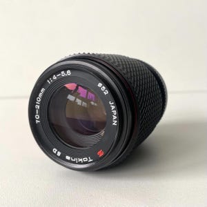 Könnte beinhalten: Ein schwarzes Tokina SD 70-210mm 1:4-5.6 Kameraobjektiv mit einem Filterdurchmesser von 52mm. Das Objektiv verfügt über einen strukturierten Gummigriffring, ein Frontglaselement mit violetten und orangen Reflexionen sowie weiße Beschriftungen.
