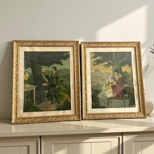May include: Two framed vintage prints with gold ornate frames. The prints depict scenes with figures in a landscape setting. One print shows two men, and the other shows a couple. The prints are displayed on a white cabinet.