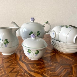 May include: A white porcelain tea set with a grapevine design. The set includes a teapot, creamer, sugar bowl, and four teacups and saucers. The set is decorated with green leaves and purple grapes.