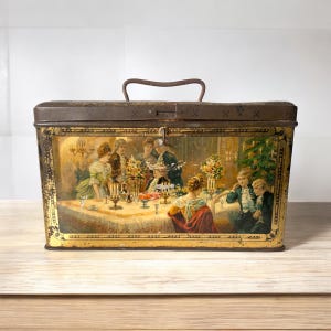 May include: Vintage metal tin box with a hinged lid and a handle. The box features a colourful illustration of people gathered around a table with candles and flowers. The illustration is set against a gold background.
