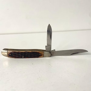 May include: A vintage pocket knife with two open blades. The handle is brown with a textured pattern. The blades are silver and the knife is open, displaying the blades at different angles. The knife is on a white surface.